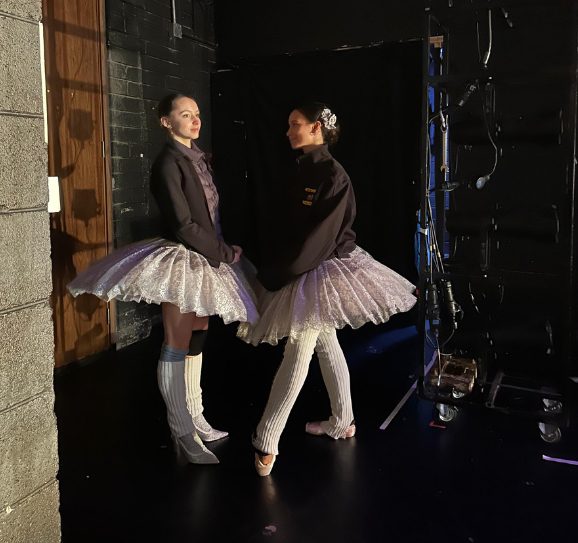 Two ballerinas in white tutus and striped leg warmers pose backstage, smiling in warm light beside theater equipment.